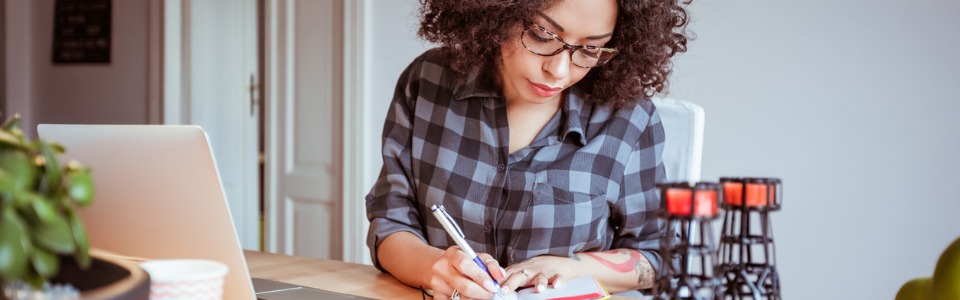 Junge Frau mit Brille sitzt am Schreibtisch vor dem Laptop und schreibt in ihr Notizbuch