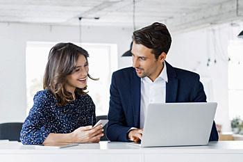 Mann und Frau sitzen vor Laptop am Schreibtisch.